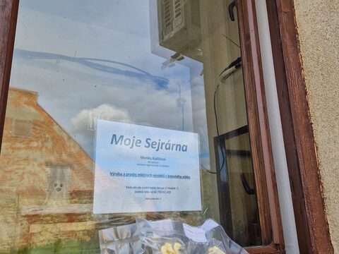 Výdejní okénko sýrárny zvenku, s cedulí a balením sejrovek na parapetu
